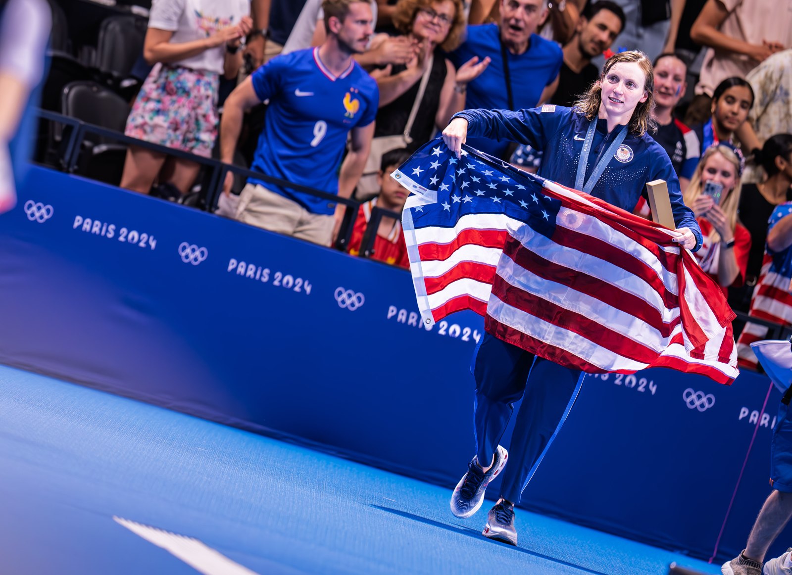 Katie Ledecky carries an American flag beside the pool at the Paris 2024 Olympics.